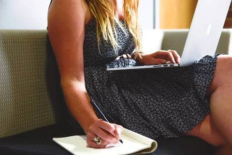 Girl sitting on coach with apple laptop on lap and taking notes on the side